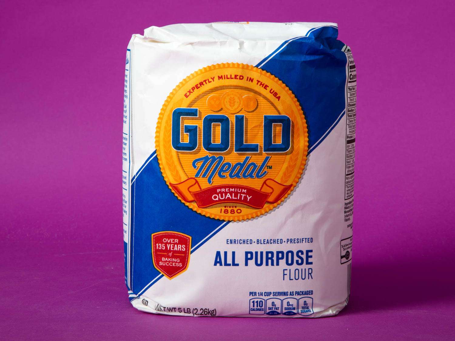 A bag of Gold Medal Blue Label bleached all-purpose flour against a purple background.
