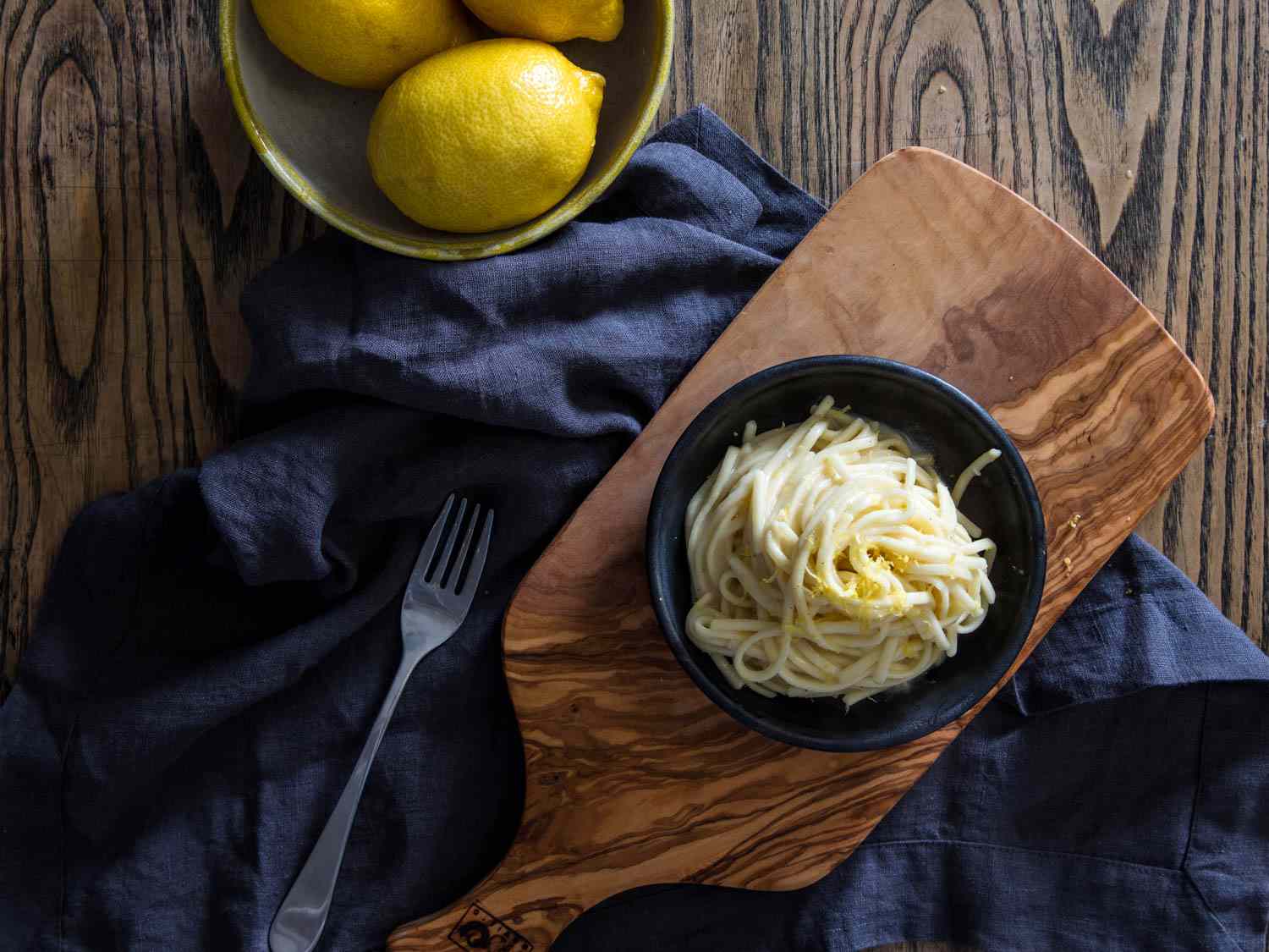 A black bowl of pasta on a wooden board, with a fork alongside and a bowl of lemons.