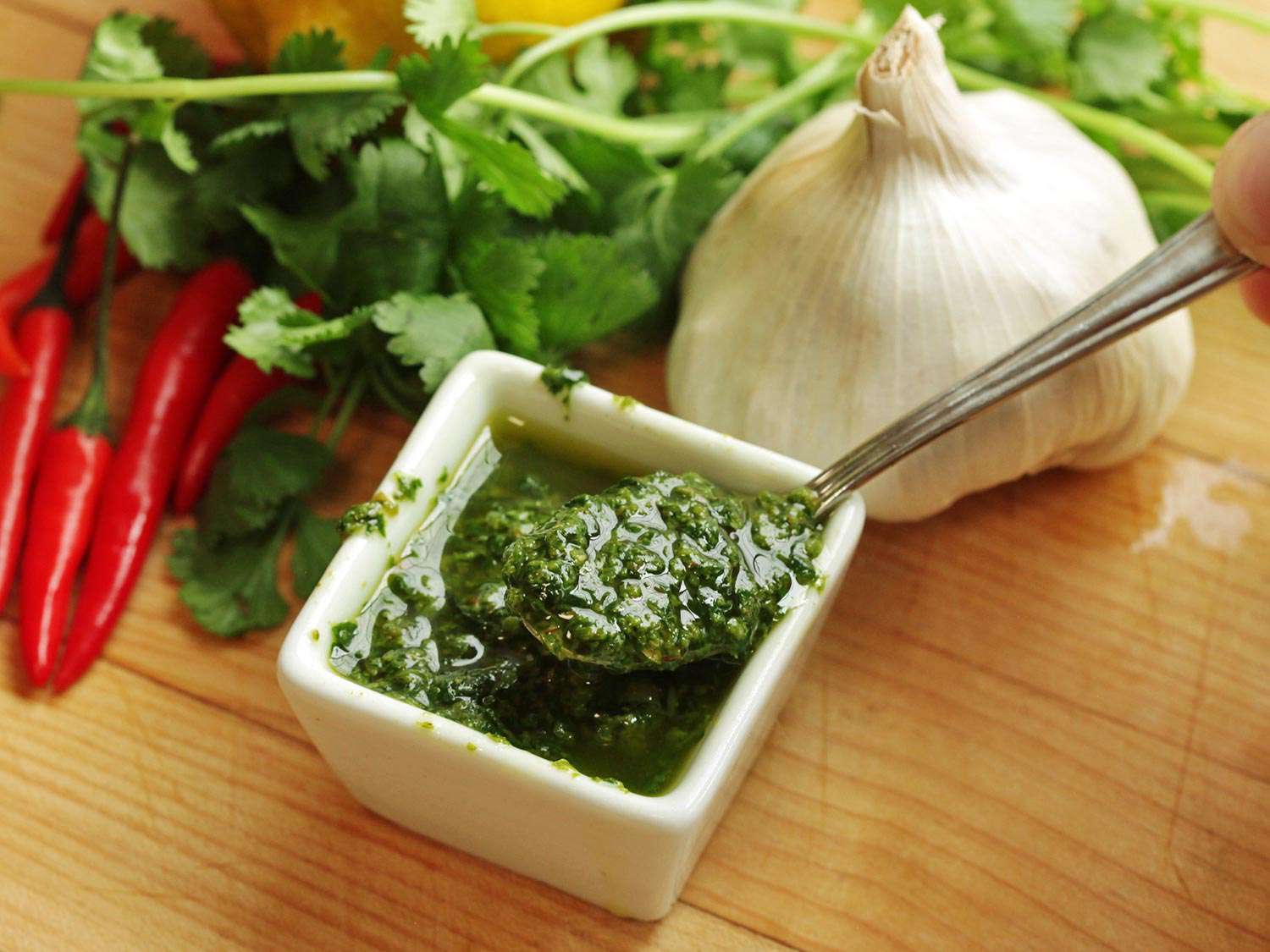A square dish of bright-green zhug (Yemenite hot sauce) next to a head of garlic, peppers, and herbs