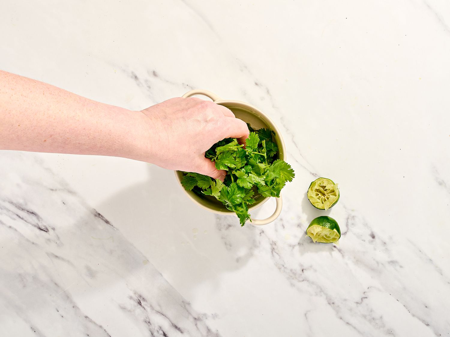 A hand placing cilantro in a bowl next to a halved lime on a marble surface
