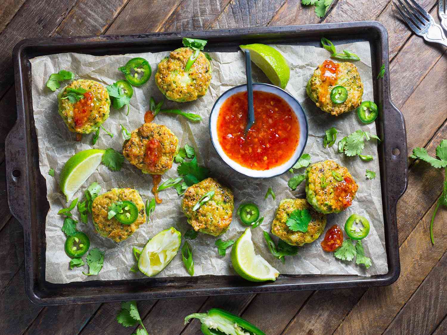 A baking sheet of Thai-style shrimp cakes with sweet chili dipping sauce and lime wedges.