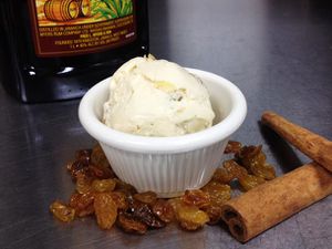 A scoop of rum raisin ice cream served in a ramekin. A bottle of dark rum, golden raisins, and cinnamon sticks are nearby.