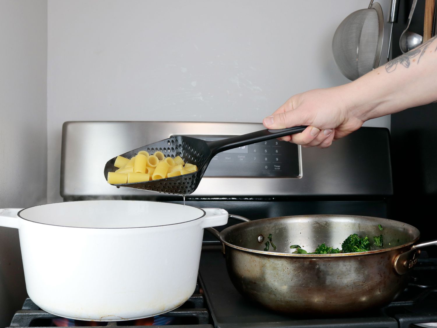A person using a colander spoon to scoop pasta out of a Dutch oven