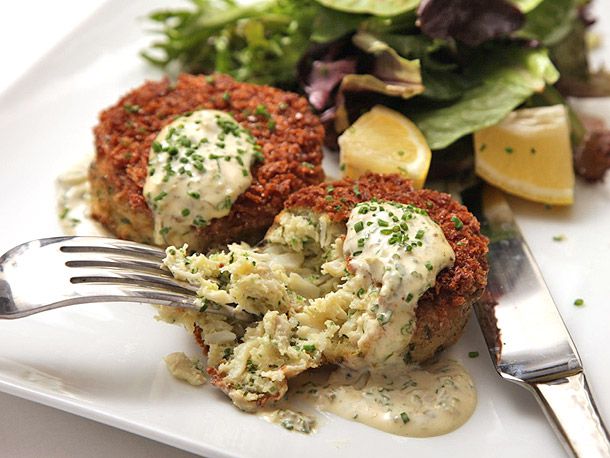 A crab cake being split open by a fork and a knife resting on a white plate. 