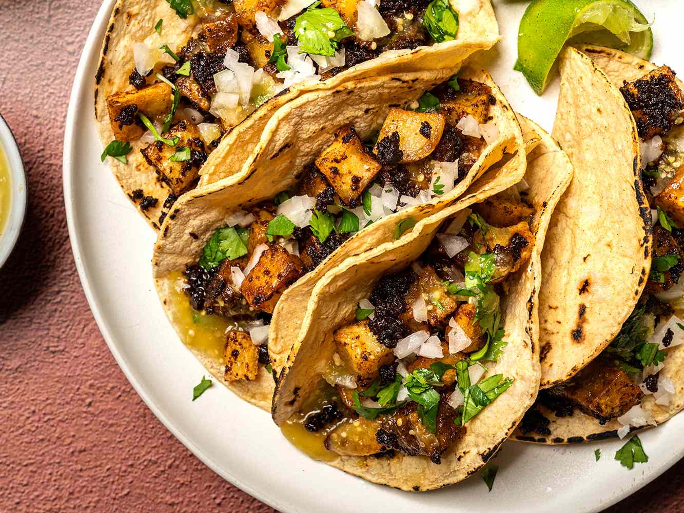 Three tacos filled with chorizo and potatoes garnished with onions and cilantro served on a plate with lime wedges