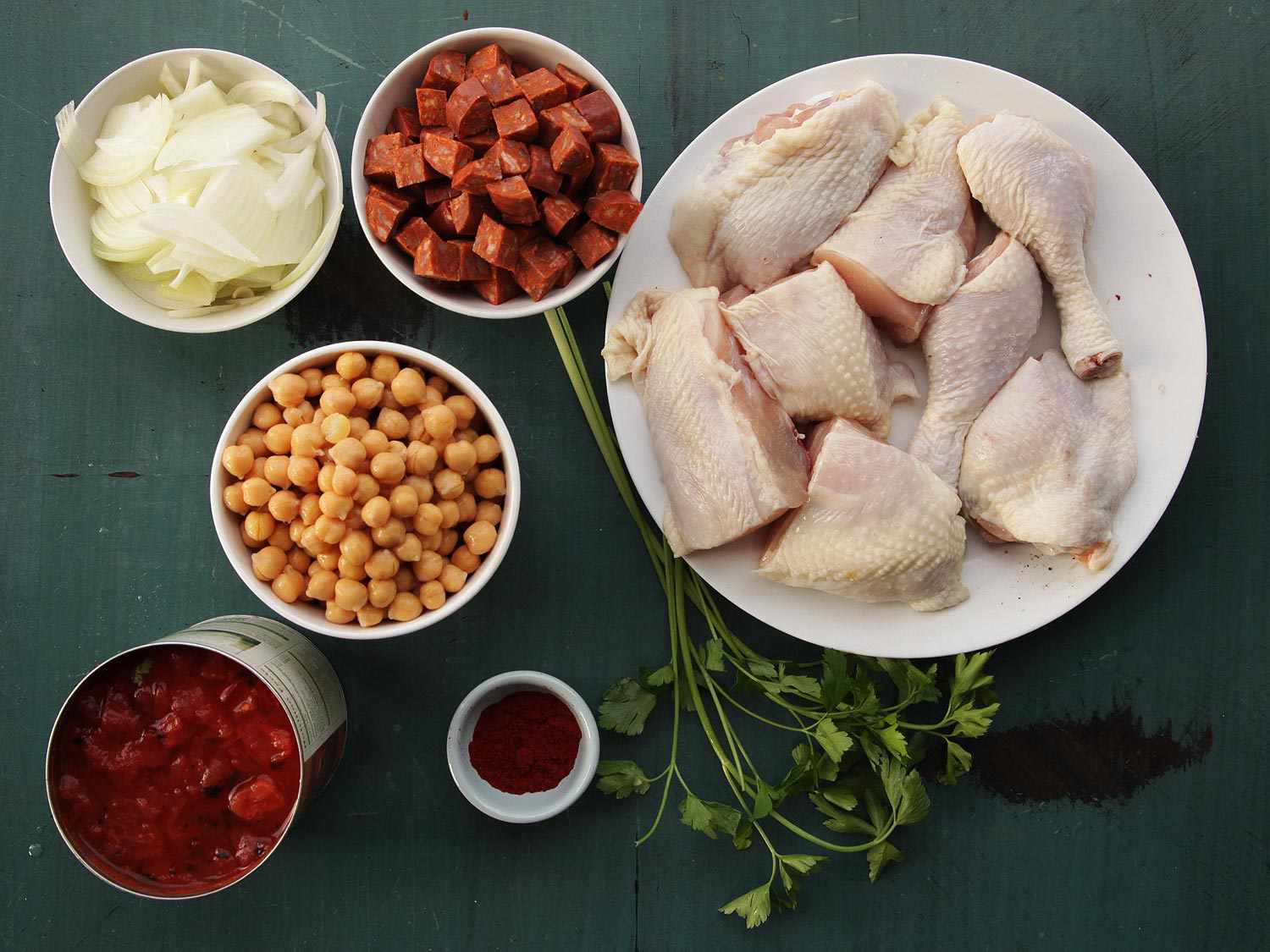 Overhead view of the prepped ingredients for the recipe: chicken parts, chorizo, chickpeas, onion, canned tomatoes, parsley, paprika.