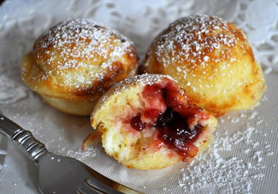 Three ebelskivers on a white cloth, dusted with powdered sugar. One is torn is cut in half, showing the lingonberry jam filling.