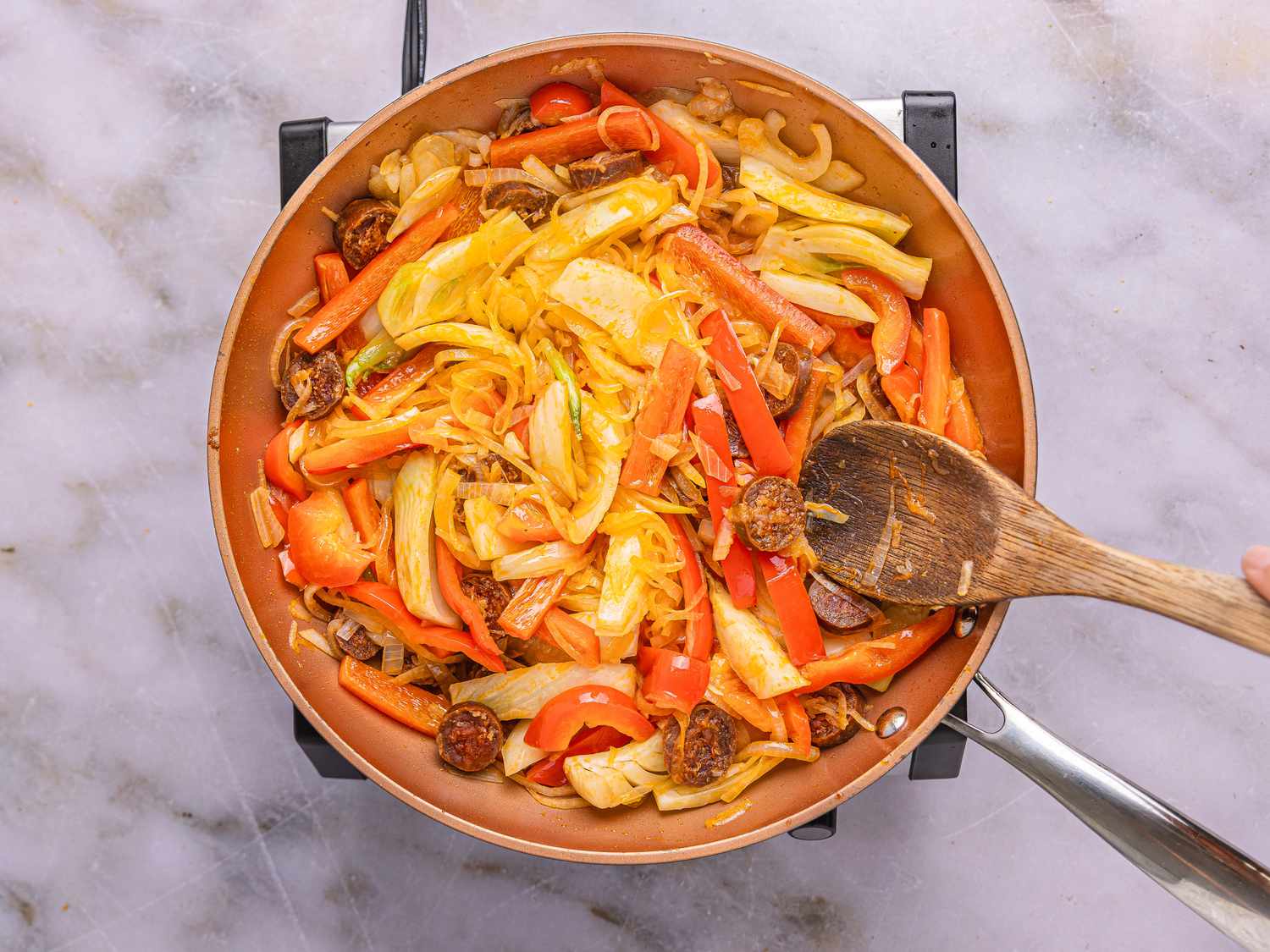 A skillet containing cooked vegetables and sliced chorizo, being stirred with a wooden spoon