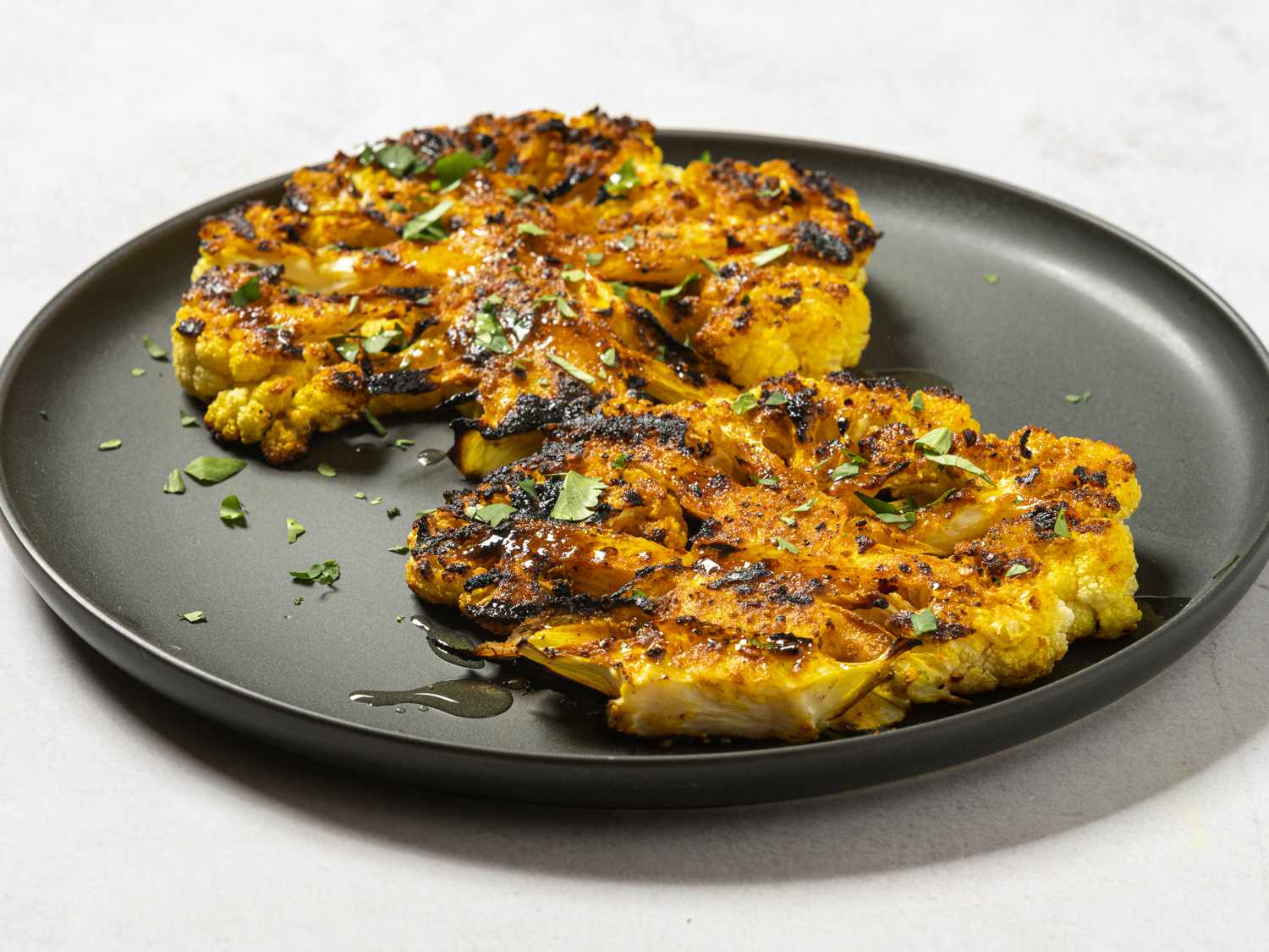 Slices of grilled spiced cauliflower on a black ceramic plate, drizzled with olive oil and sprinkled with chopped herbs.