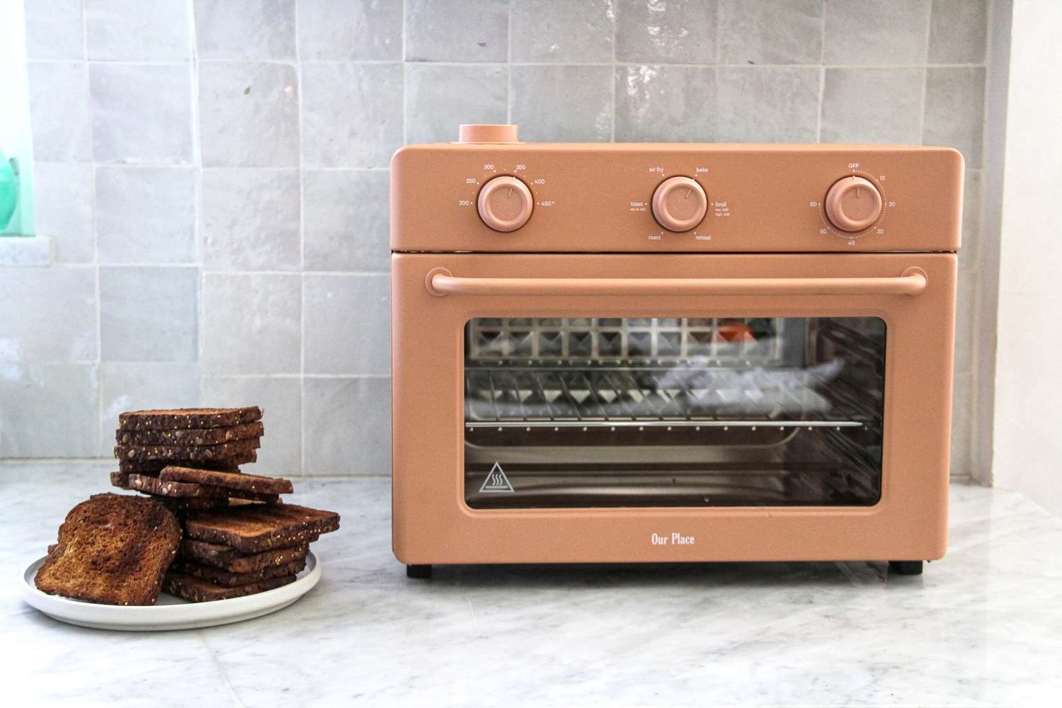 An Our Place Wonder Oven beside a plate of toasted bread slices on a kitchen counter, with a neutral backsplash in the background