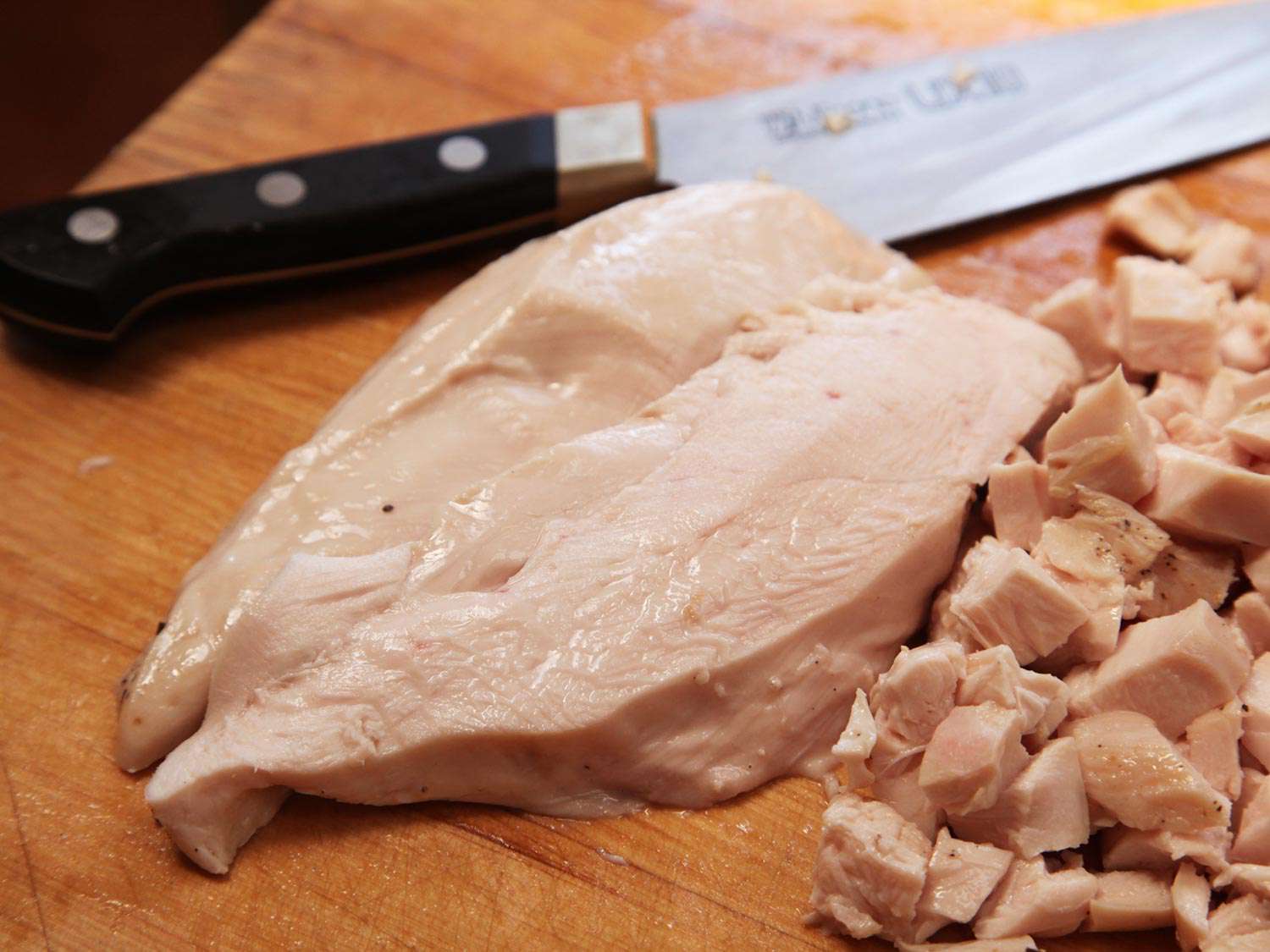 Chicken breast on a wooden surface, next to a pile of diced chicken and a chef's knife.