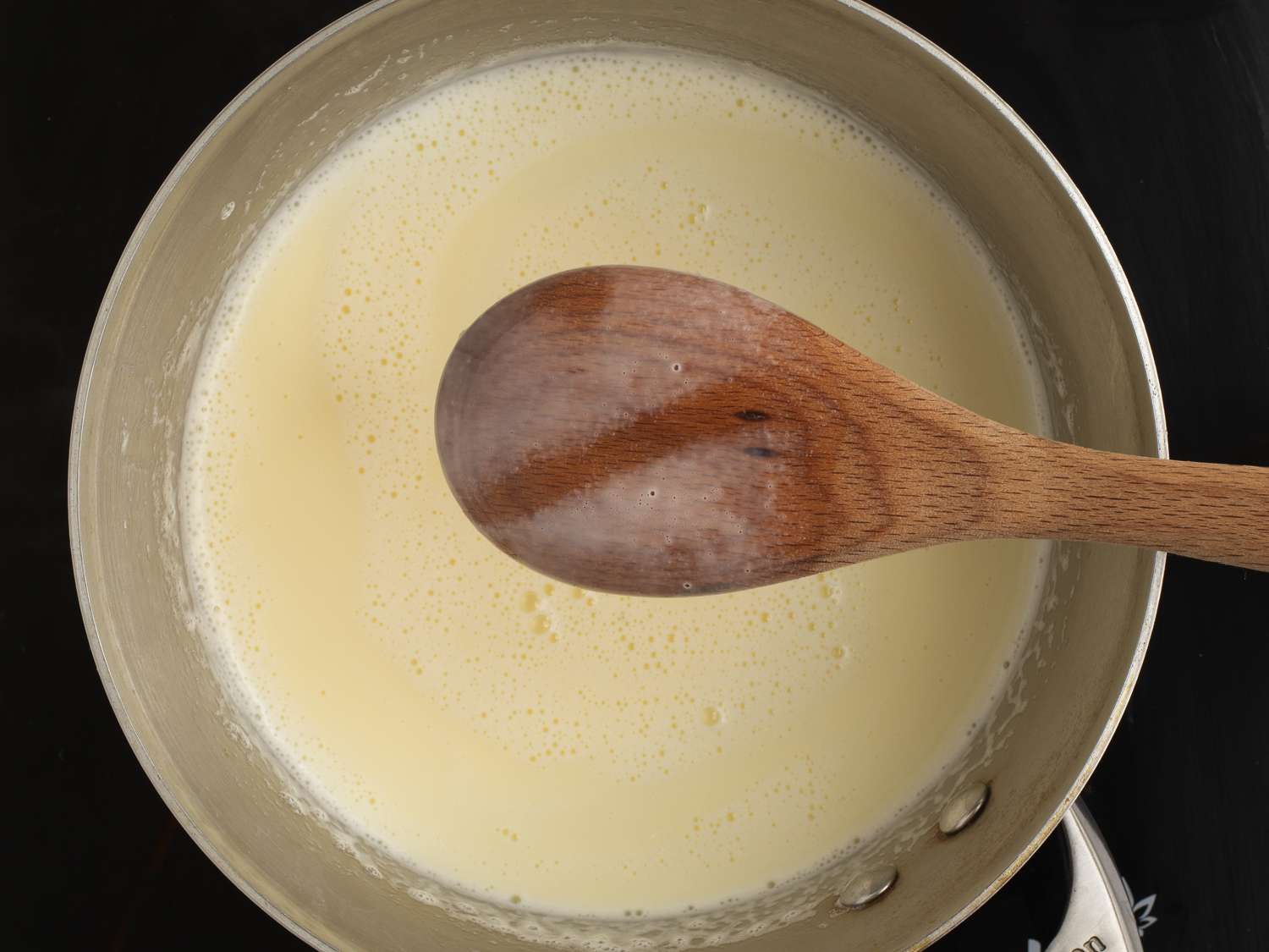 A wooden spoon held above a saucepan holding the combined custard. A line has been drawn through the cream mixture coating the back of the spoon, showing it's reached the correct consistency.