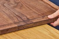 A hand holding the edge of a Virginia Boys Kitchens Walnut Wood Cutting Board resting on a table