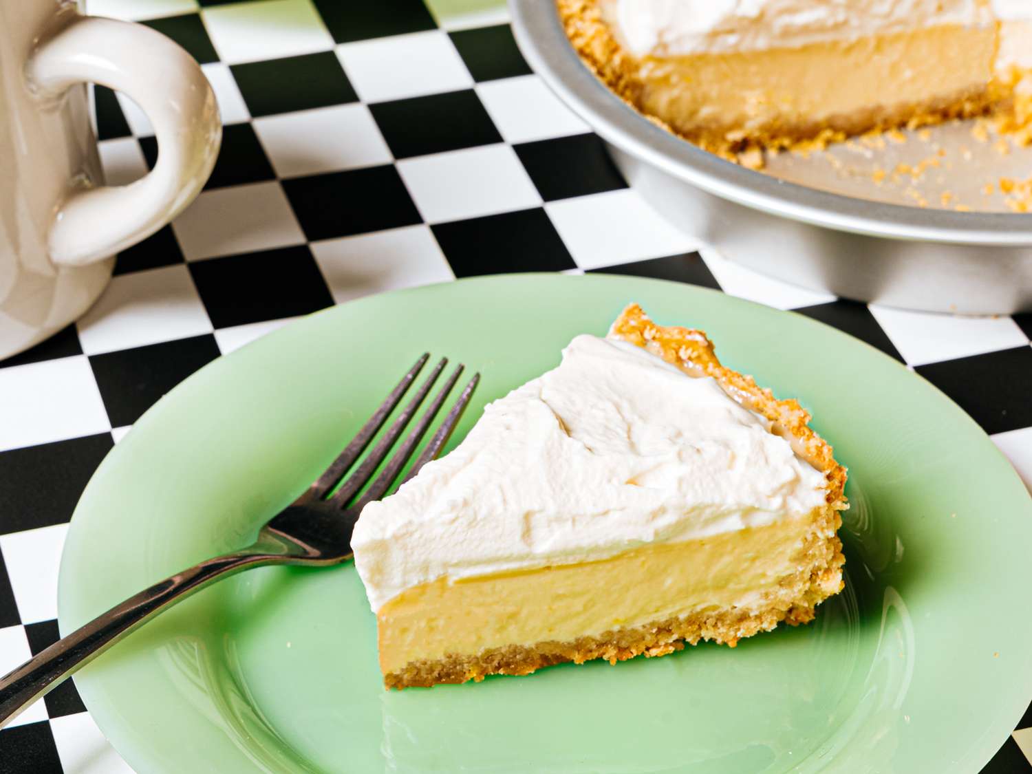 A slice of creamtopped pie on a green plate with a fork a pie plate with the rest of the pie in the background