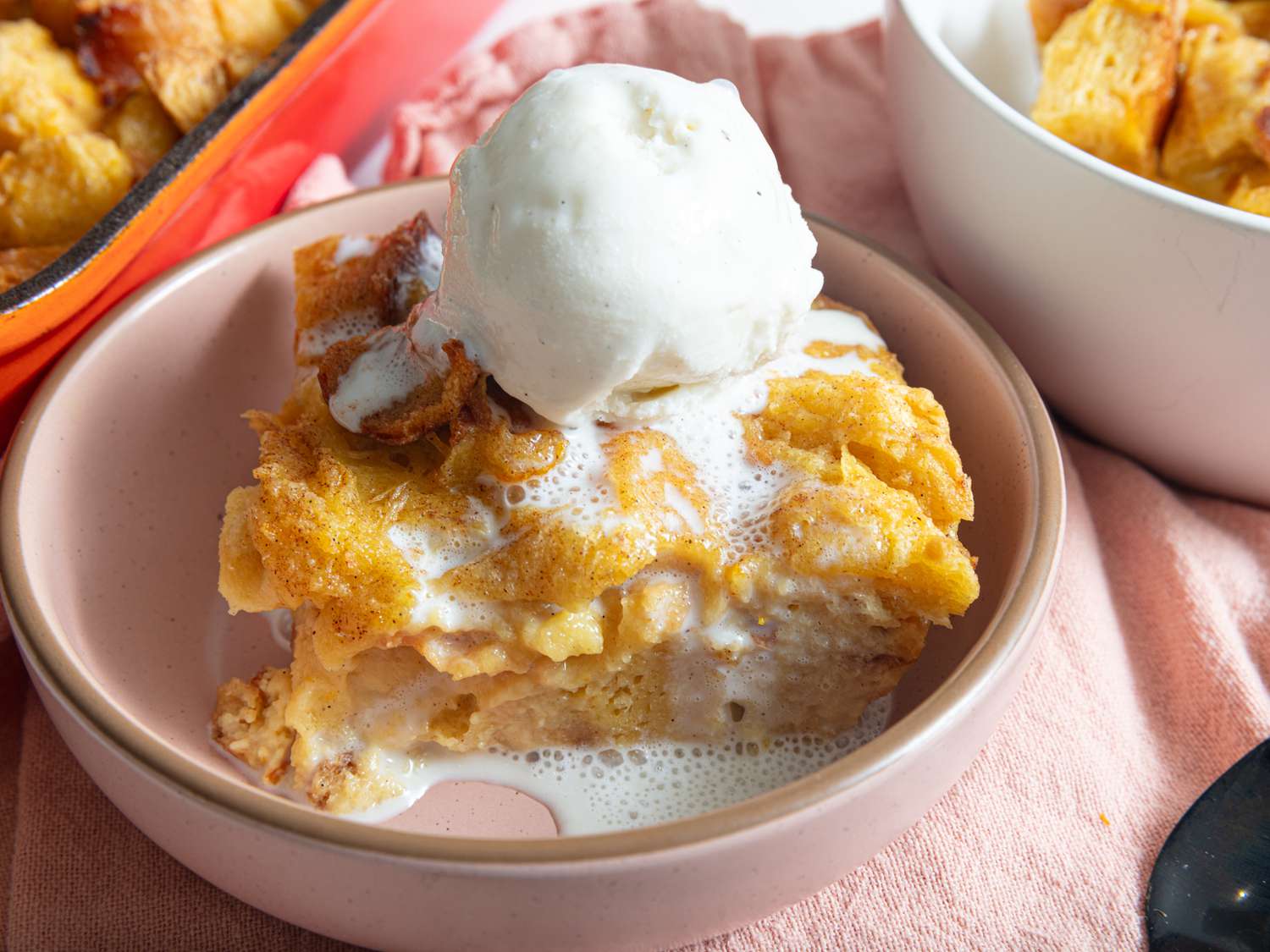 Soft pink bowl with bread pudding a la mode, on a soft pink textile, with a white bowl of bread pudding and full red tray of bread pudding on the sides. 