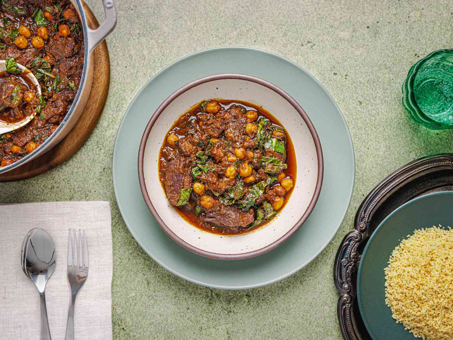 bowl of beef stew on a blue plate, with pot of stew to one side, plate of rice to another side, and cutlery. All on a green stone surface. 