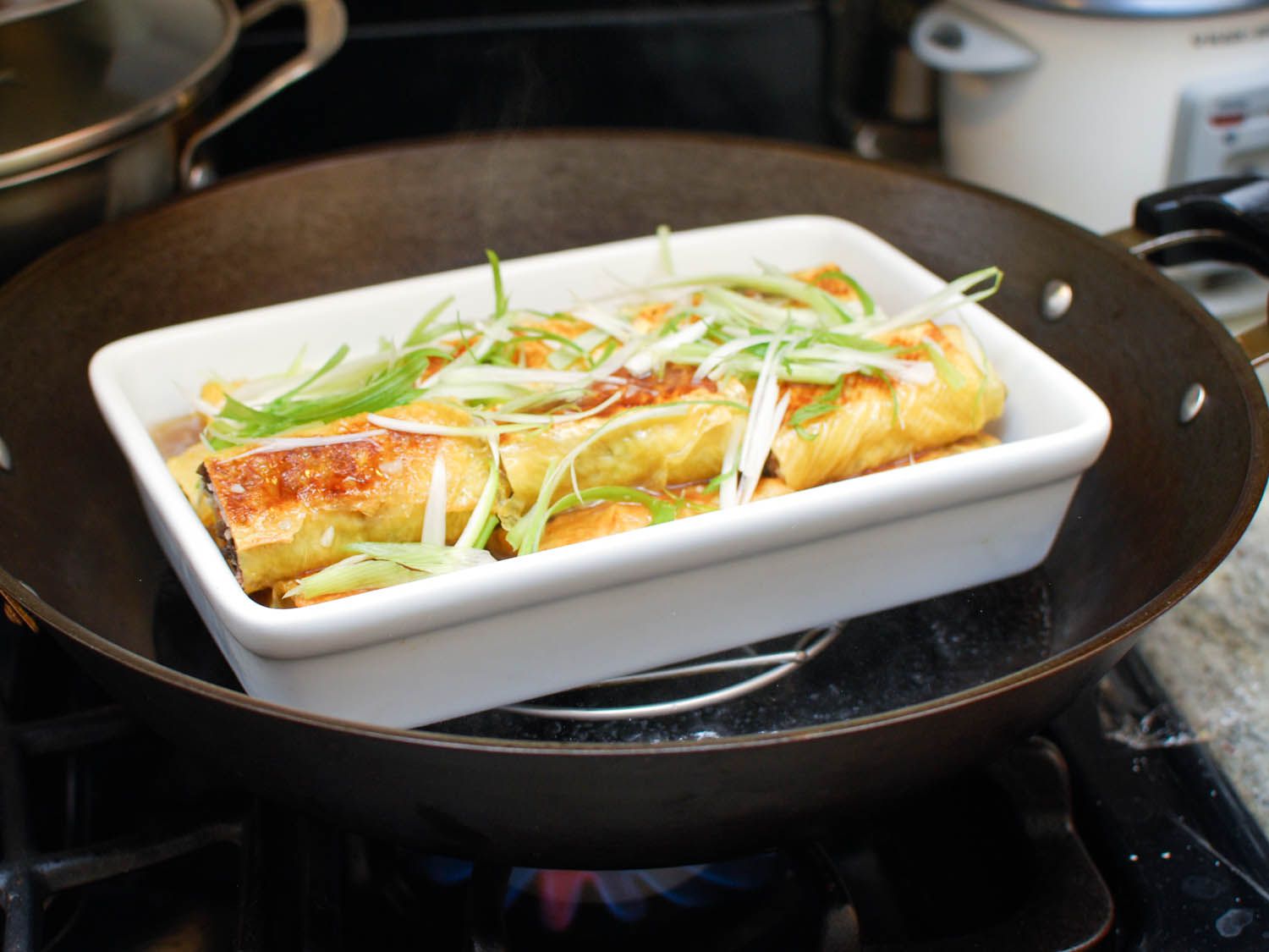 A baking dish full of the bean curd rolls, thickened stock, and sliced scallion is placed on a rack inside a wok, ready for steaming.