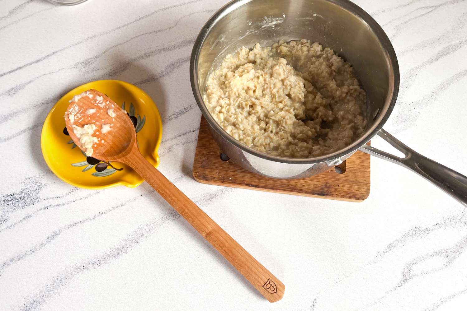A pot of oatmeal on a wooden trivet with a Field Company Cherry Wood Spoon resting on a yellow dish nearby