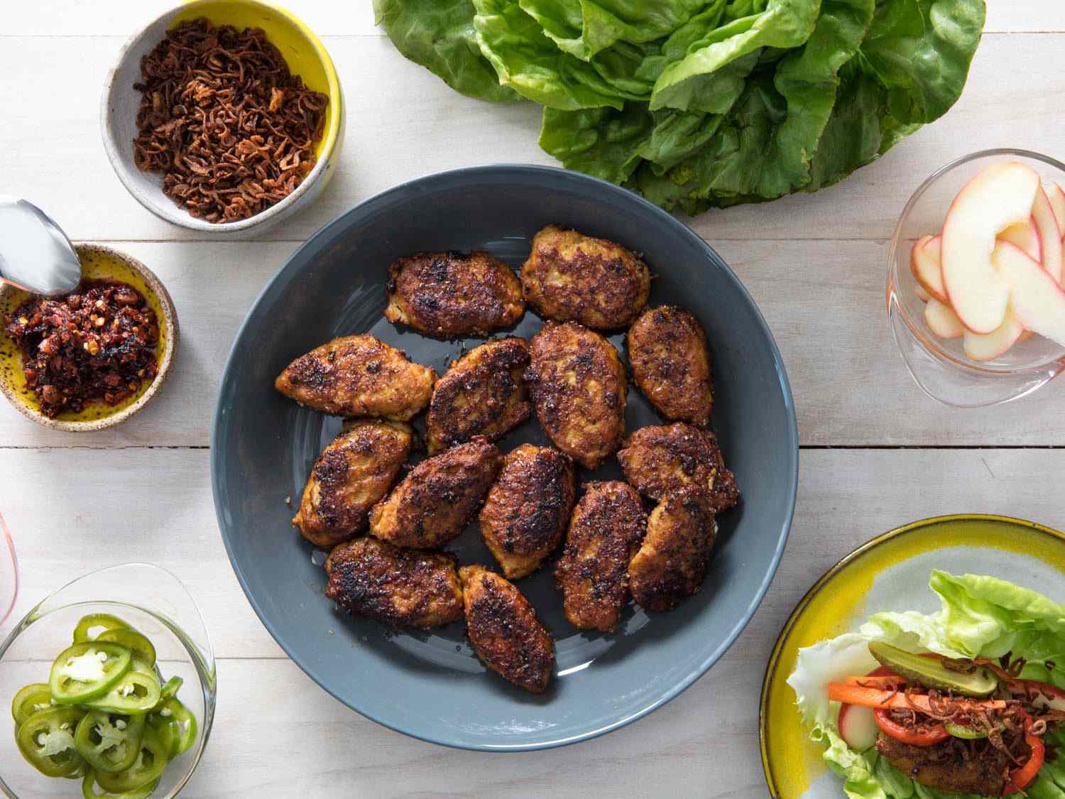 Overhead view of the finished meatballs served on a table with lettuce leaves, crispy shallots, chile crisp, sliced chiles, and apple slices.