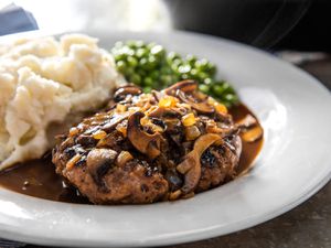 A Salisbury steak smothered in mushroom gravy with mashed potatoes and peas