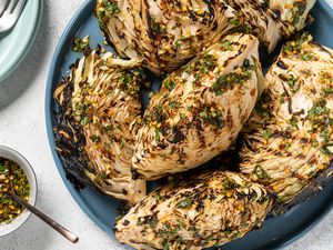 Grilled cabbage wedges drizzled with sauce inside of a blue platter. On the lefthand side of the image are a small bowl with a metal spoon holding additional sauce, and in the top left corner is a blue plate holding silverware.