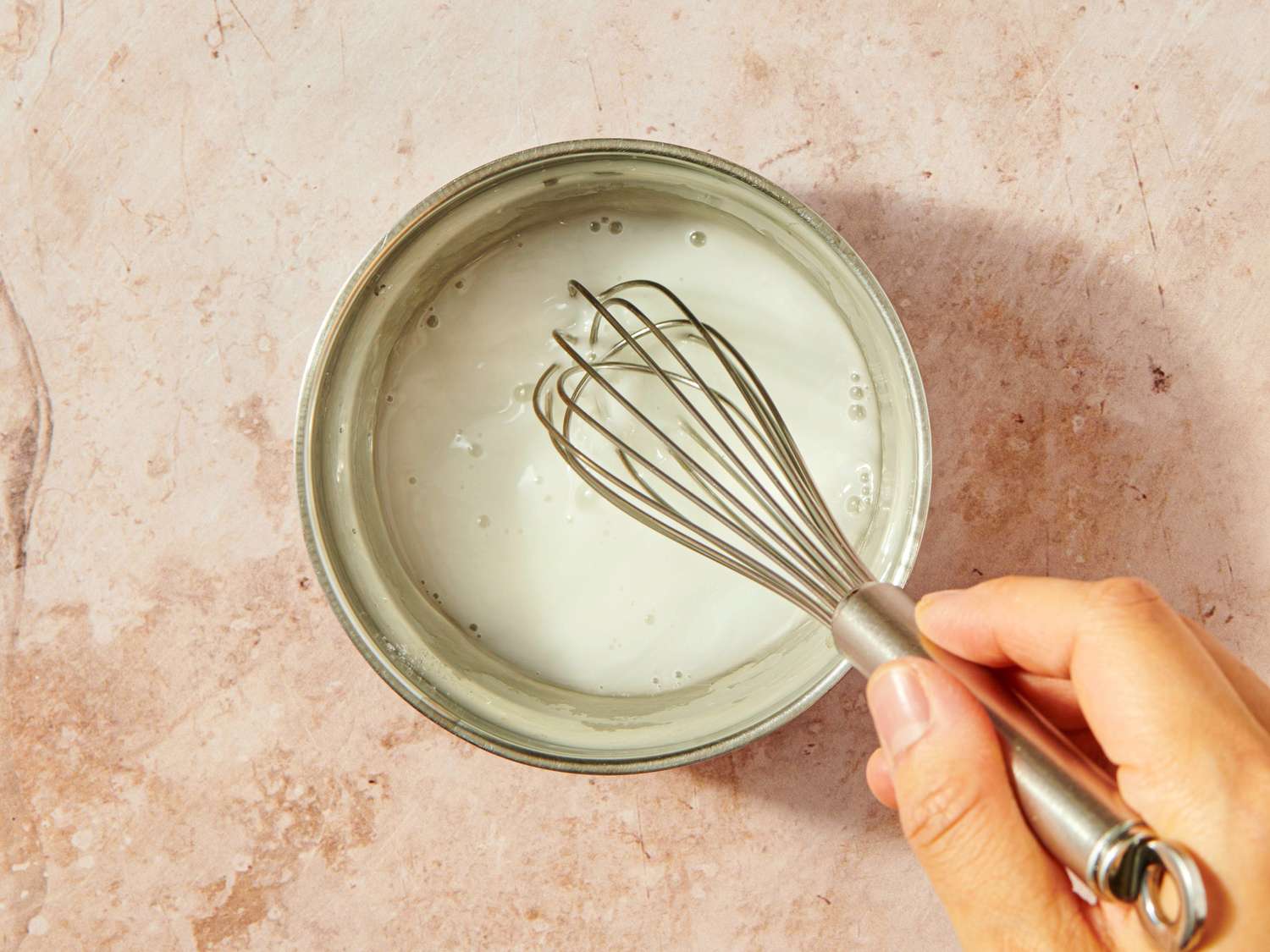 A hand holding a whisk stirring a white liquid in a metallic bowl on a light surface