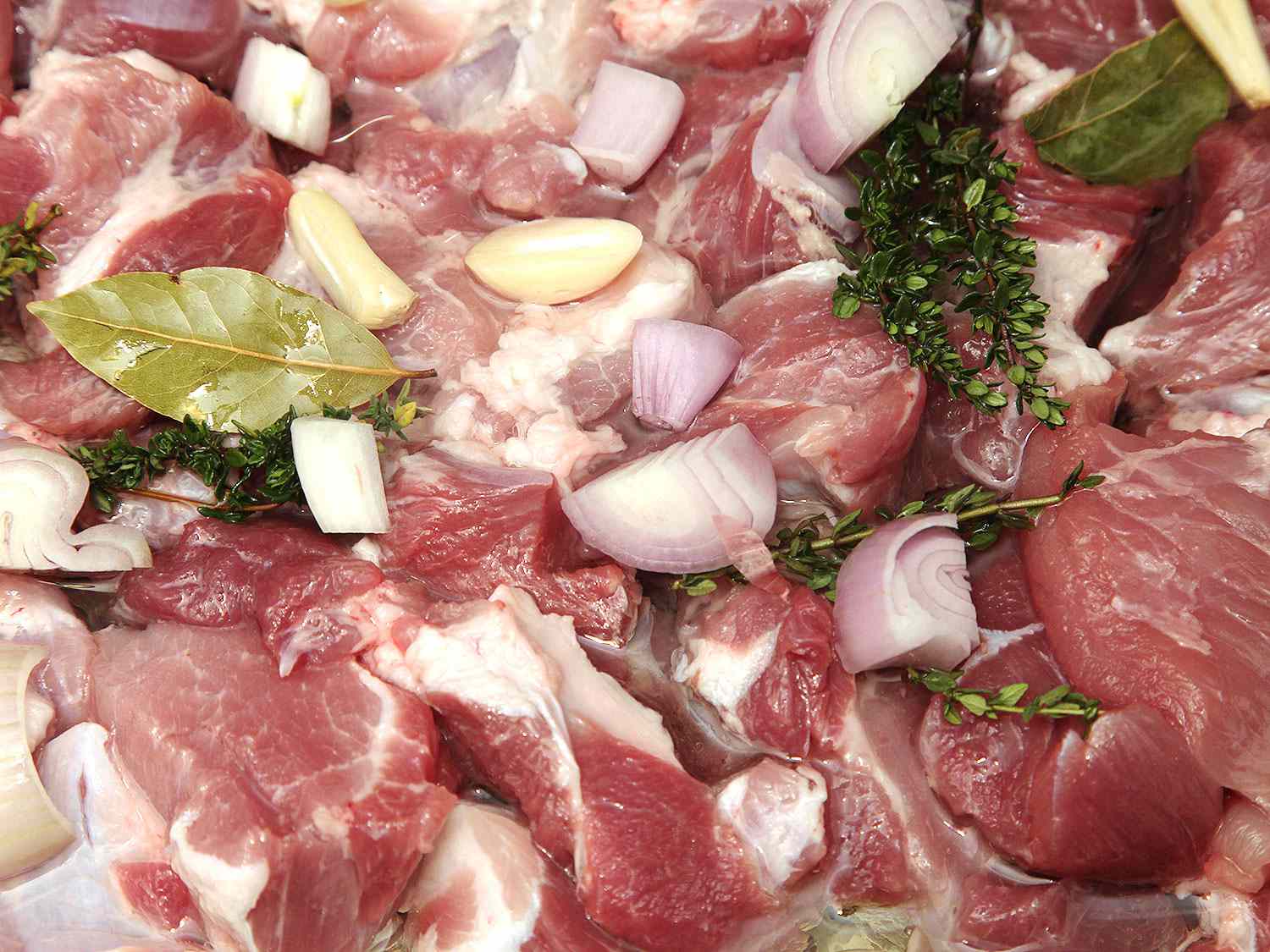 Closeup of raw pork shoulder chunks, smothered with garlic, shallot, bay leaves, thyme, and oil.