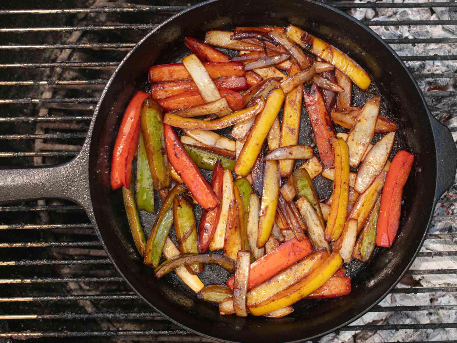 Peppers and onions cooking in a cast iron skillet on a charcoal grill.