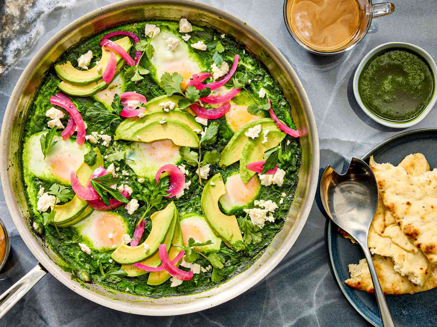 Pot of shakshuka on grey stone surface, with coffee cup, and side of pita. 