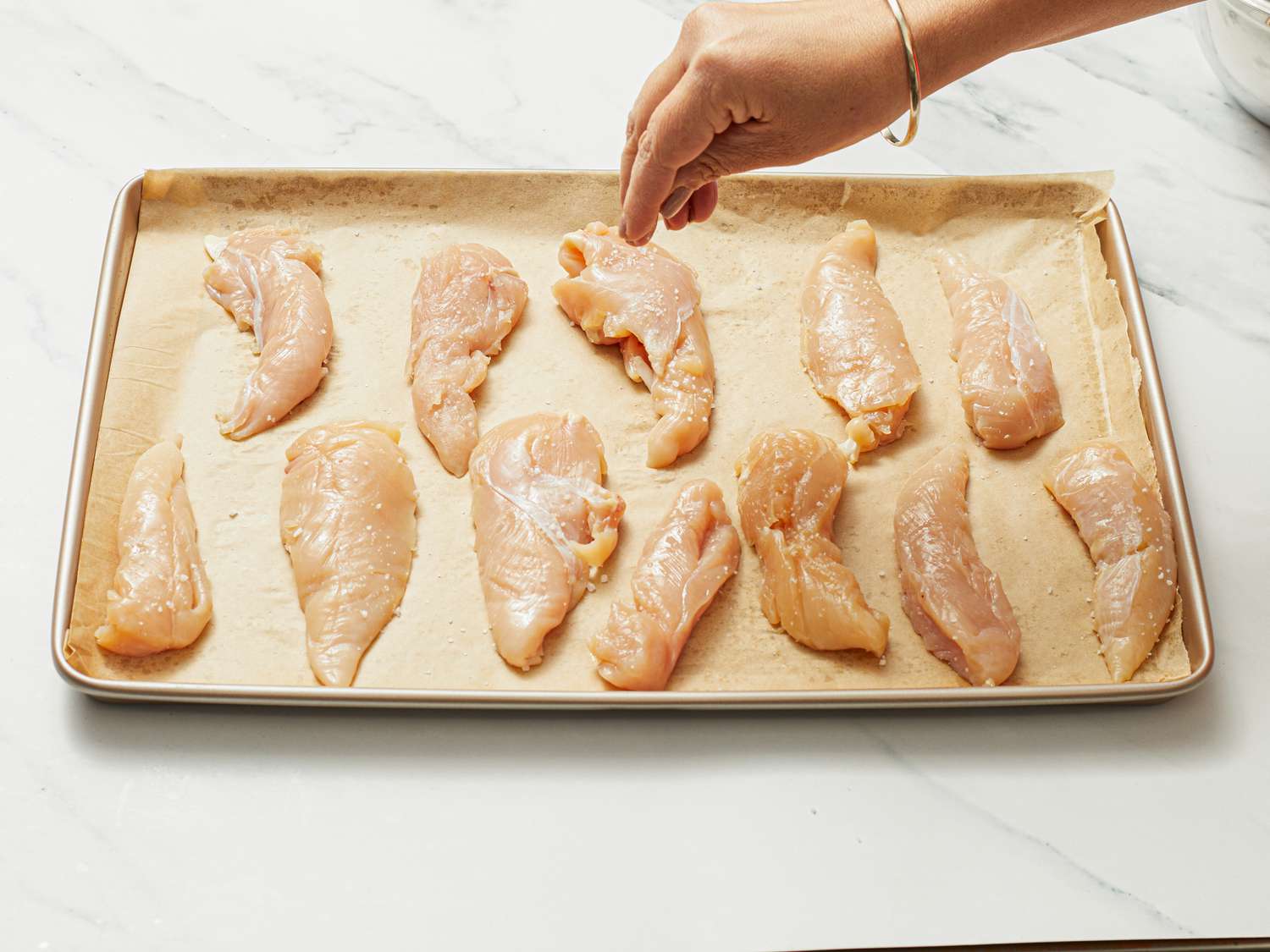 Chicken tenders arranged on a baking sheet, a hand sprinkling seasoning over them.