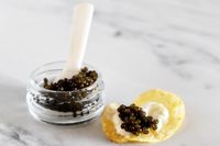 An open jar of Olma kaluga royal caviar next to a potato chip topped with crème fraîche and a scoop of caviar