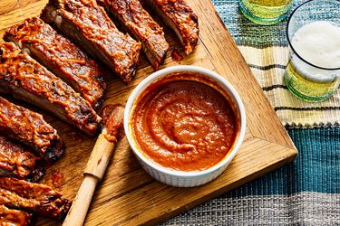 overhead view of bbq sauce on a cutting board alongside ribs and glasses of beer