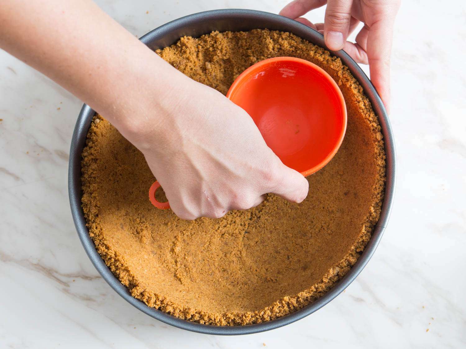 Hand using a dry measuring cup to press cookie crumb crust into the bottom and sides of a springform pan for pumpkin cheesecake.