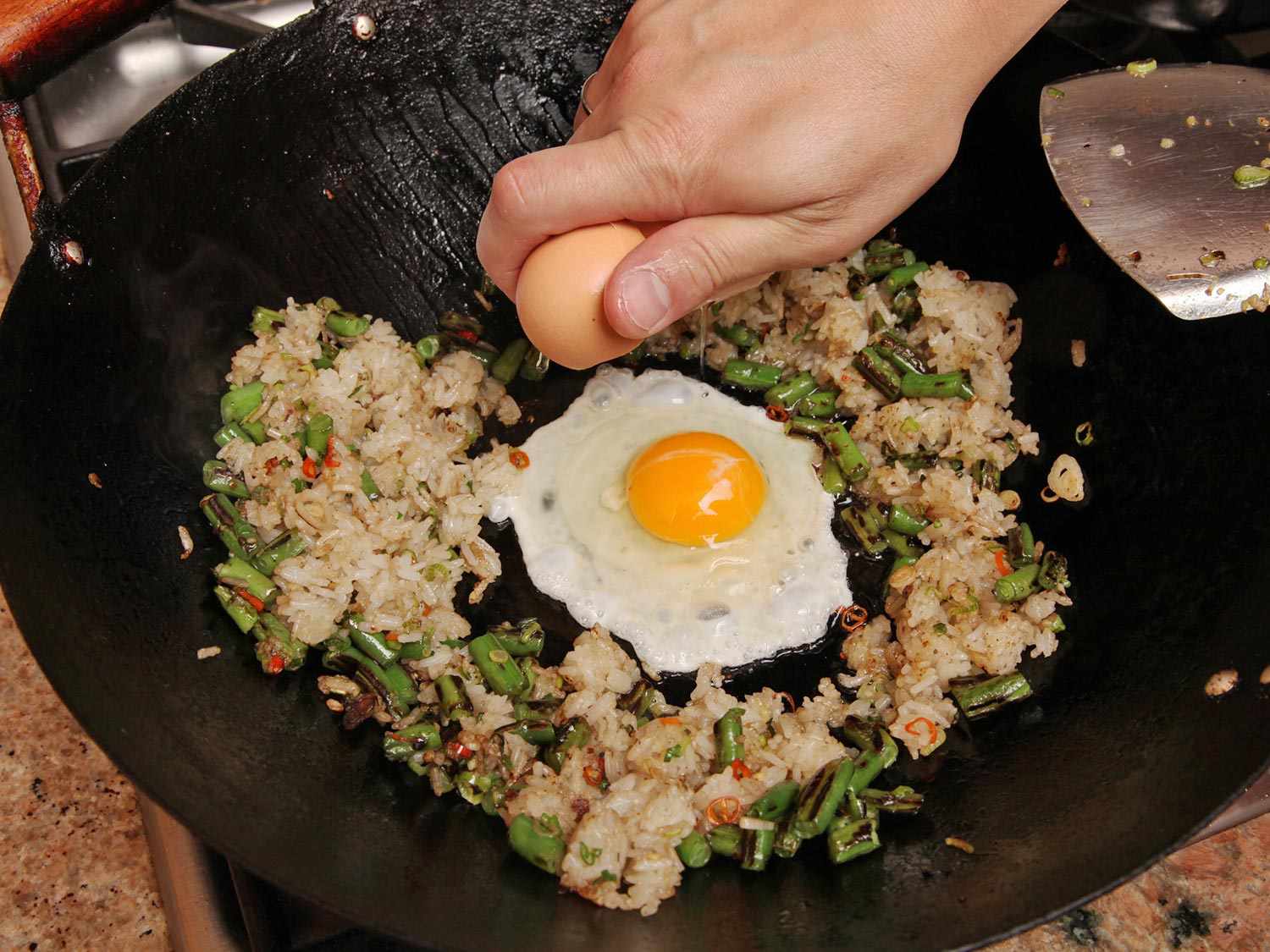 Author cracks an egg into a bare spot in the center of the wok. The fried rice and vegetables have been moved to the periphery of the pan.