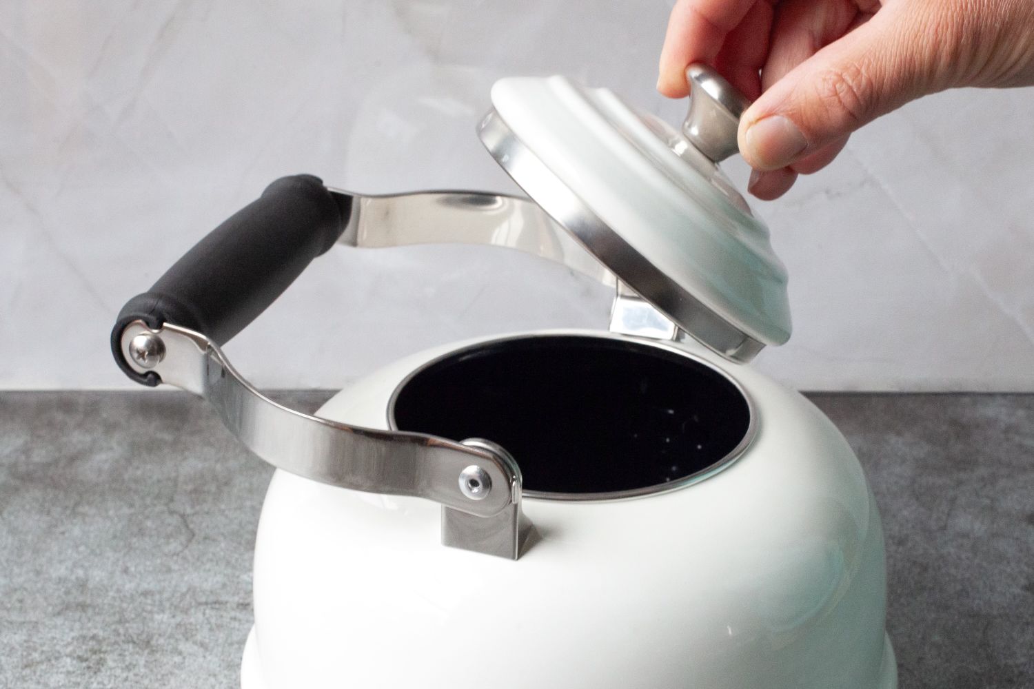 A person lifts the lid of a white teapot with a black handle and a metallic rim