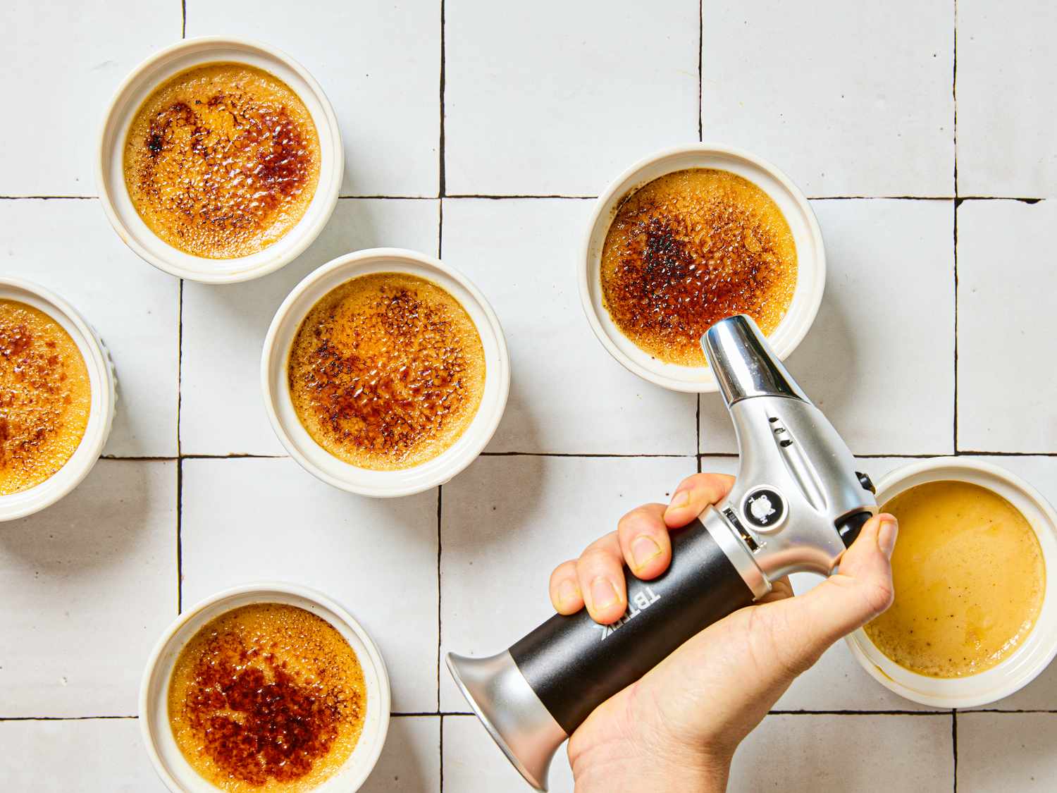 A hand using a culinary torch to caramelize the tops of crme brle desserts in ramekins