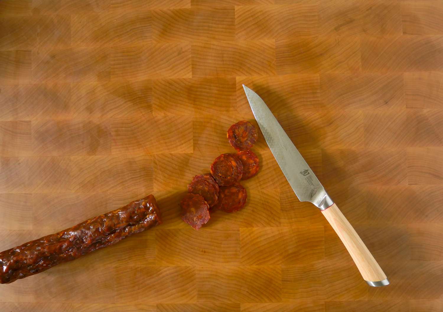 a knife on a cutting board with cut up pieces of cured sausage