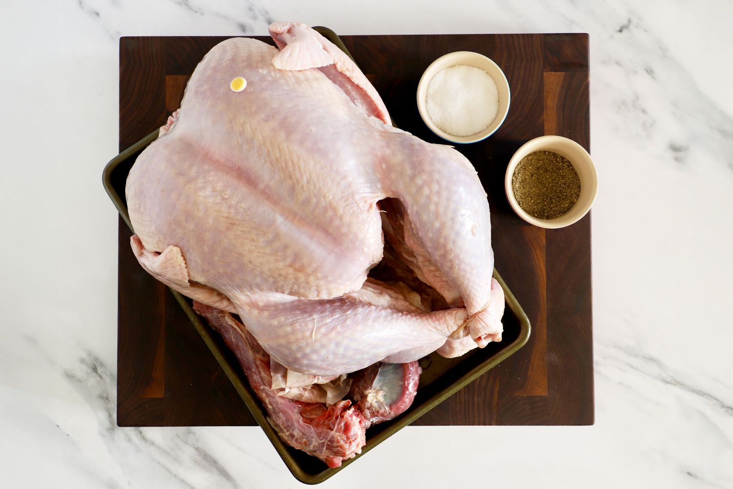 A raw whole turkey on a tray alongside bowls of salt and pepper on a wooden board
