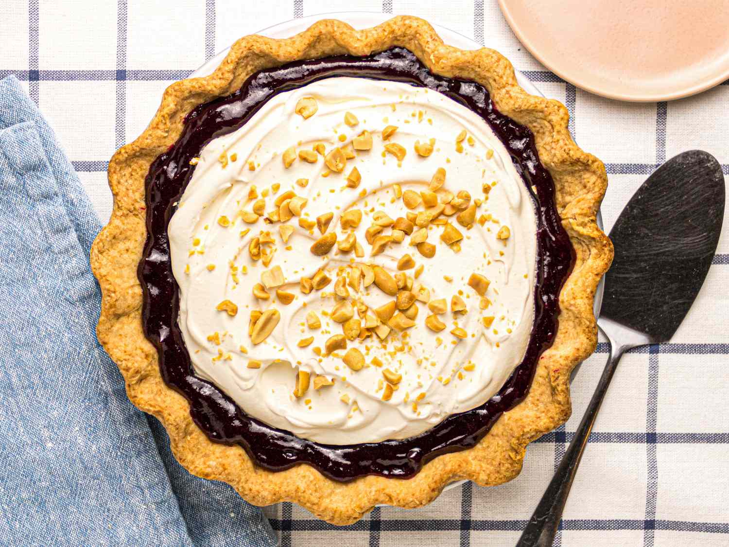 A peanut butter jelly pie topped with whipped topping and chopped peanuts on a checkered tablecloth with a pie server