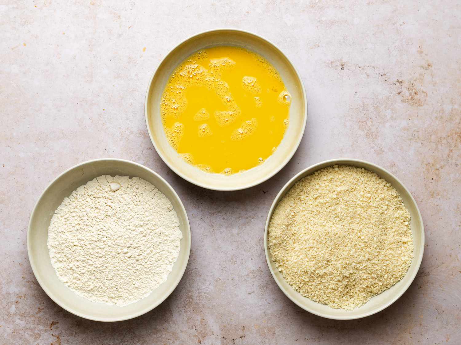 Three bowls arranged in a rough triangle. The left-most bowl contains flour, the top-center bowl contains beaten eggs, and the right-most bowl holds grated parmesan cheese mixed with panko breadcrumbs. 