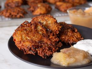 A plate of fried latkes served with applesauce and sour cream additional latkes cooling on a rack in the background