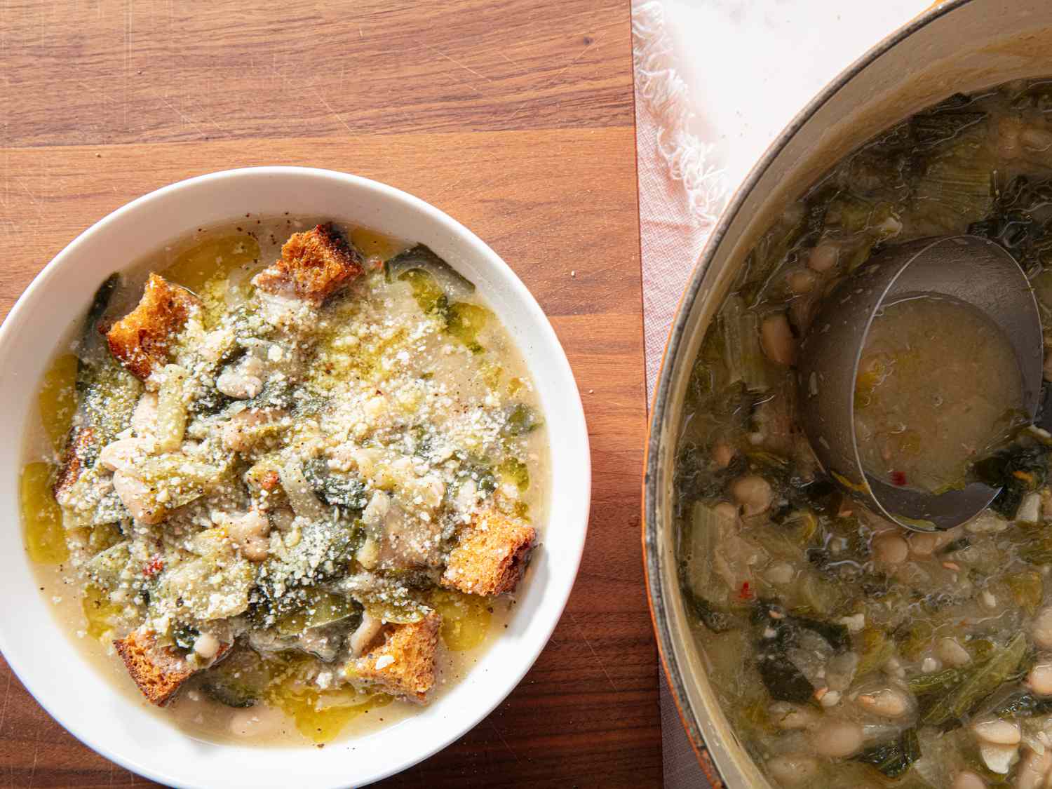 Bowl of soup with greens croutons and grated cheese next to a pot containing the same soup and a ladle