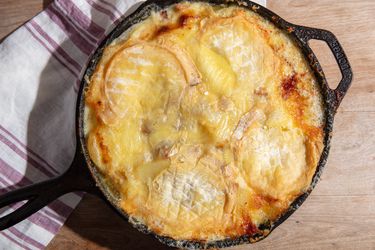 A cast iron skillet full to the brim with baked potato tartiflette, topped with melted and browned soft-rind cheese.