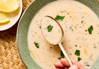 A bowl of garlic soup with garnish and a hand holding a spoon over the surface a plate of lemon slices to the side