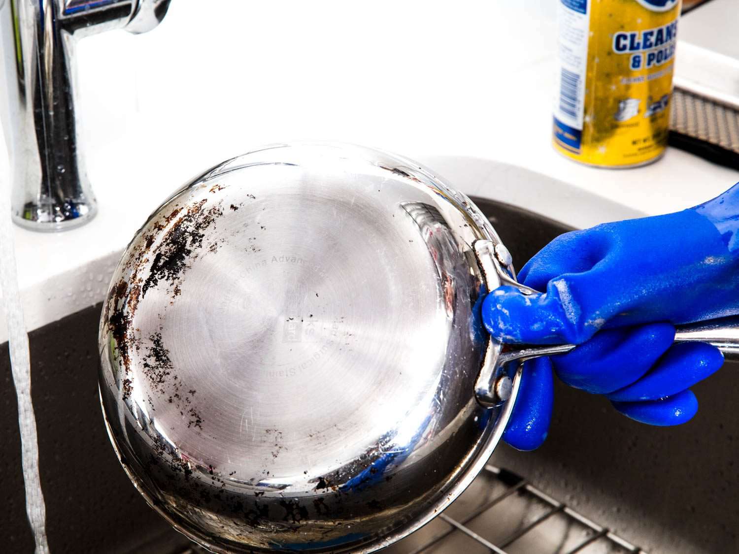 A blue-gloved hand holding a stainless steel skillet under a faucet.