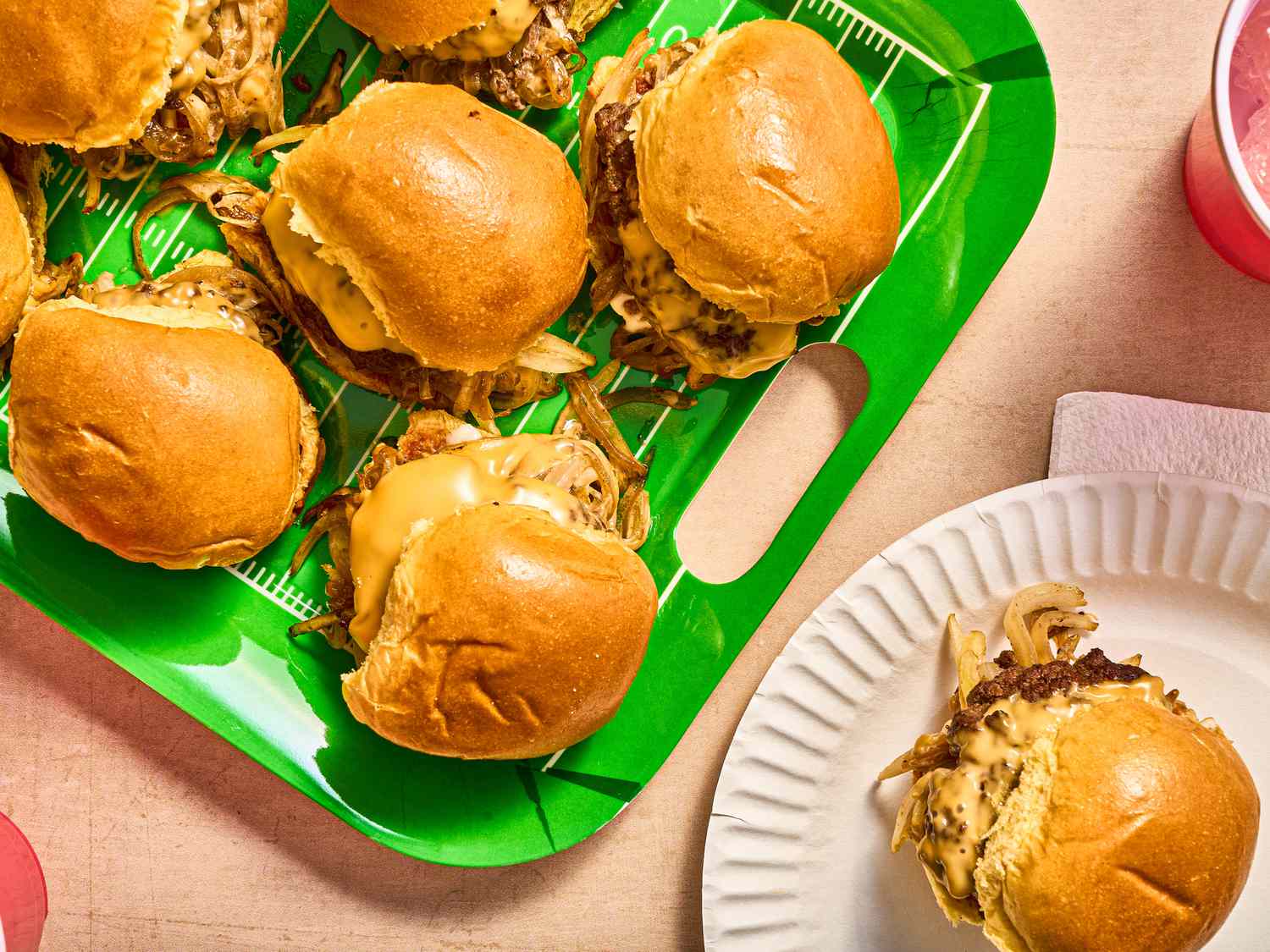 overhead angle of sliders on a bright green tray, with one slider on a paper plate in the bottom right corner, and 2 red cups on the edges of the frames