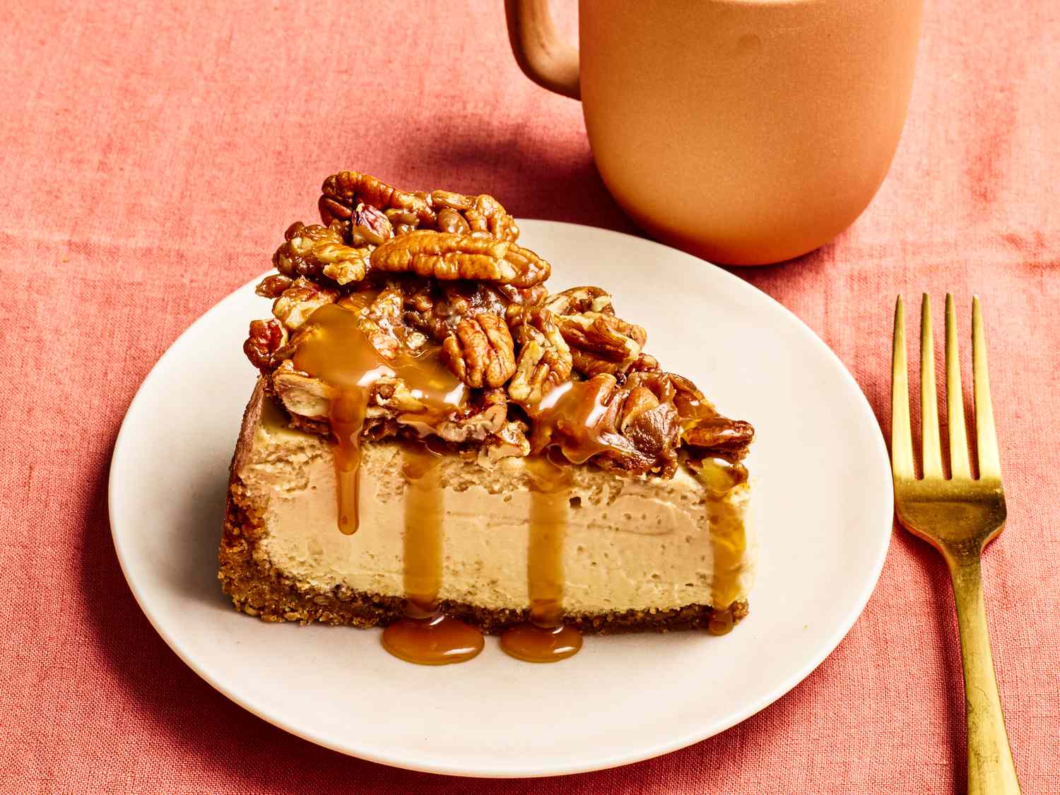 Slice of pecan pie cheesecake on a plate with a fork and mug on a table
