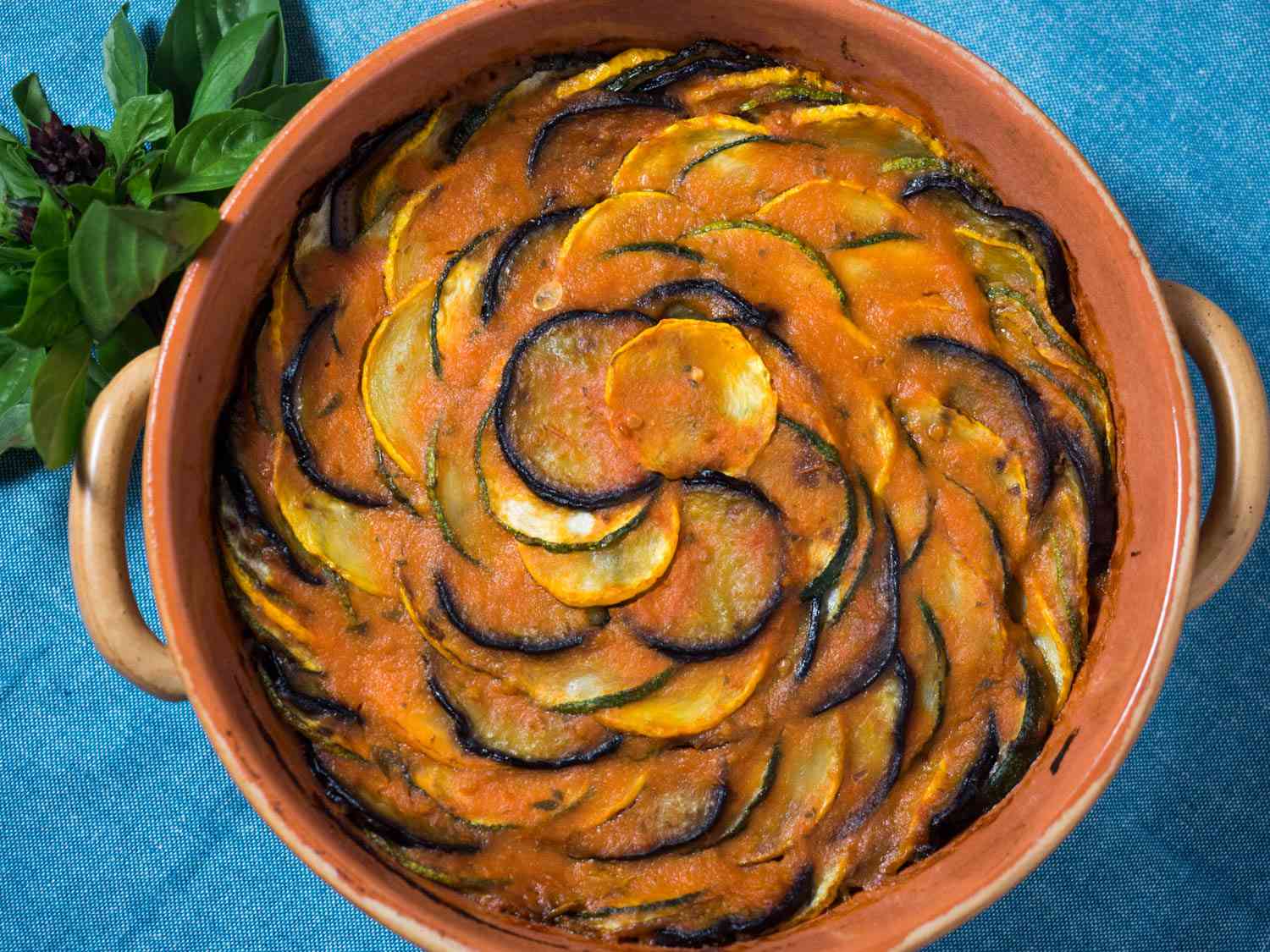 An overhead view of a pretty Provençal tian, which has concentric circles of summer squash, tomatoes, and eggplant in a crock next to fresh basil on a blue background.