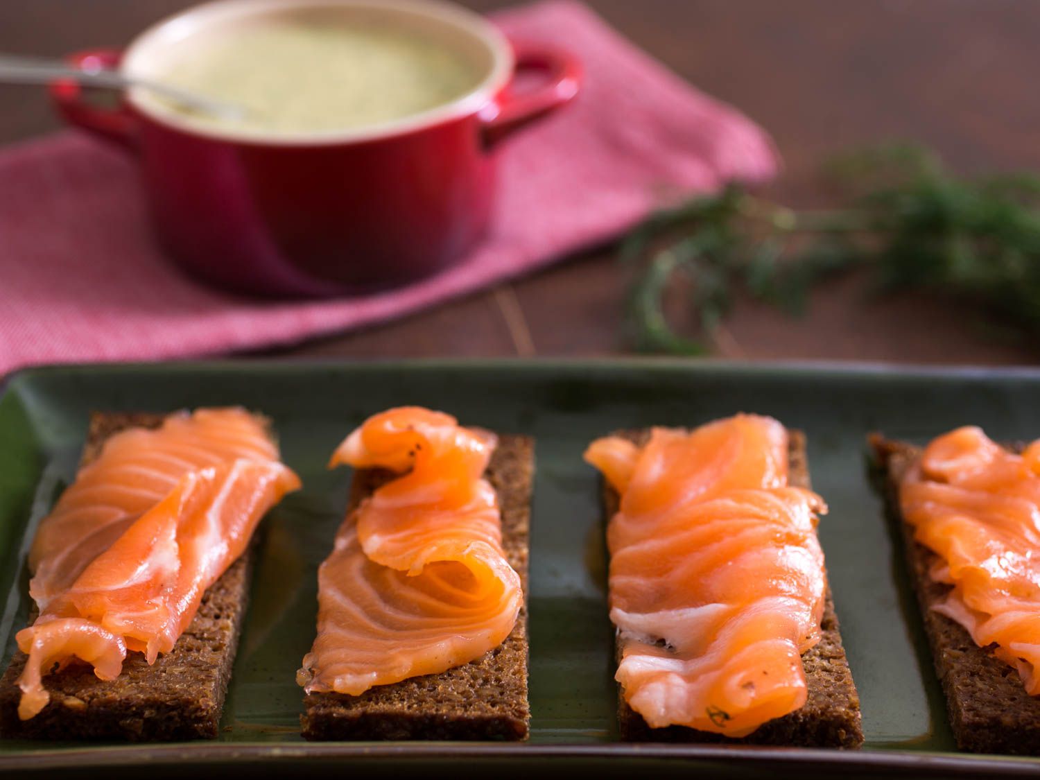 A tray of gravlax on pumpernickel slices. A bowl of hovmästarsås on the background. 