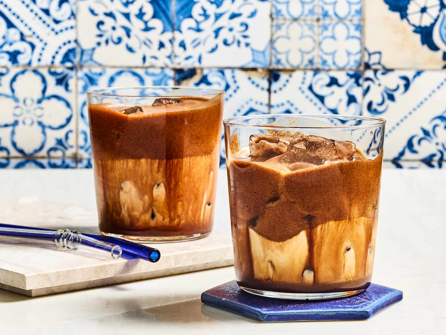 Two glasses of iced mocha beverages on a tiled background